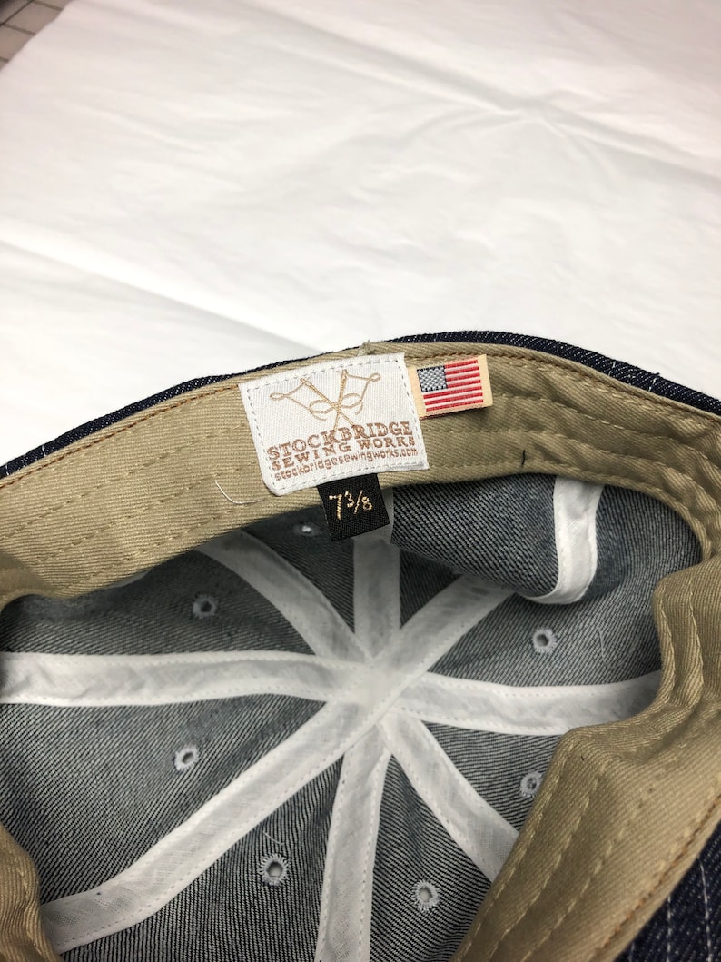 K&ouml;nnte beinhalten: Eine Baseballkappe aus Jeansstoff mit einem beigefarbenen Innenband und wei&szlig;en N&auml;hten. Die Kappe hat ein Etikett mit dem Text "STOCKBRIDGE SEWING WORKS" und eine kleine amerikanische Flagge.