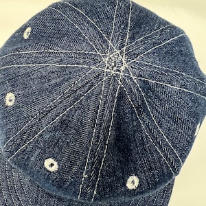 K&ouml;nnte beinhalten: Dunkelblaue Baseballkappe aus Denim mit wei&szlig;en N&auml;hten. Die Kappe hat einen gebogenen Schirm und ein sechsteiliges Design, wobei sich wei&szlig;e N&auml;hte von der Mitte der Krone ausbreiten. Kleine wei&szlig;e, gestickte Akzente sind um die Kappe platziert.