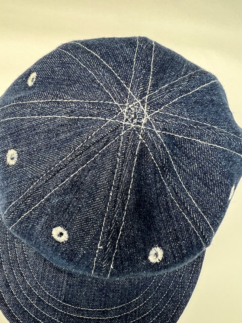 K&ouml;nnte beinhalten: Dunkelblaue Baseballkappe aus Denim mit wei&szlig;en N&auml;hten. Die Kappe hat einen gebogenen Schirm und ein sechsteiliges Design, wobei sich wei&szlig;e N&auml;hte von der Mitte der Krone ausbreiten. Kleine wei&szlig;e, gestickte Akzente sind um die Kappe platziert.