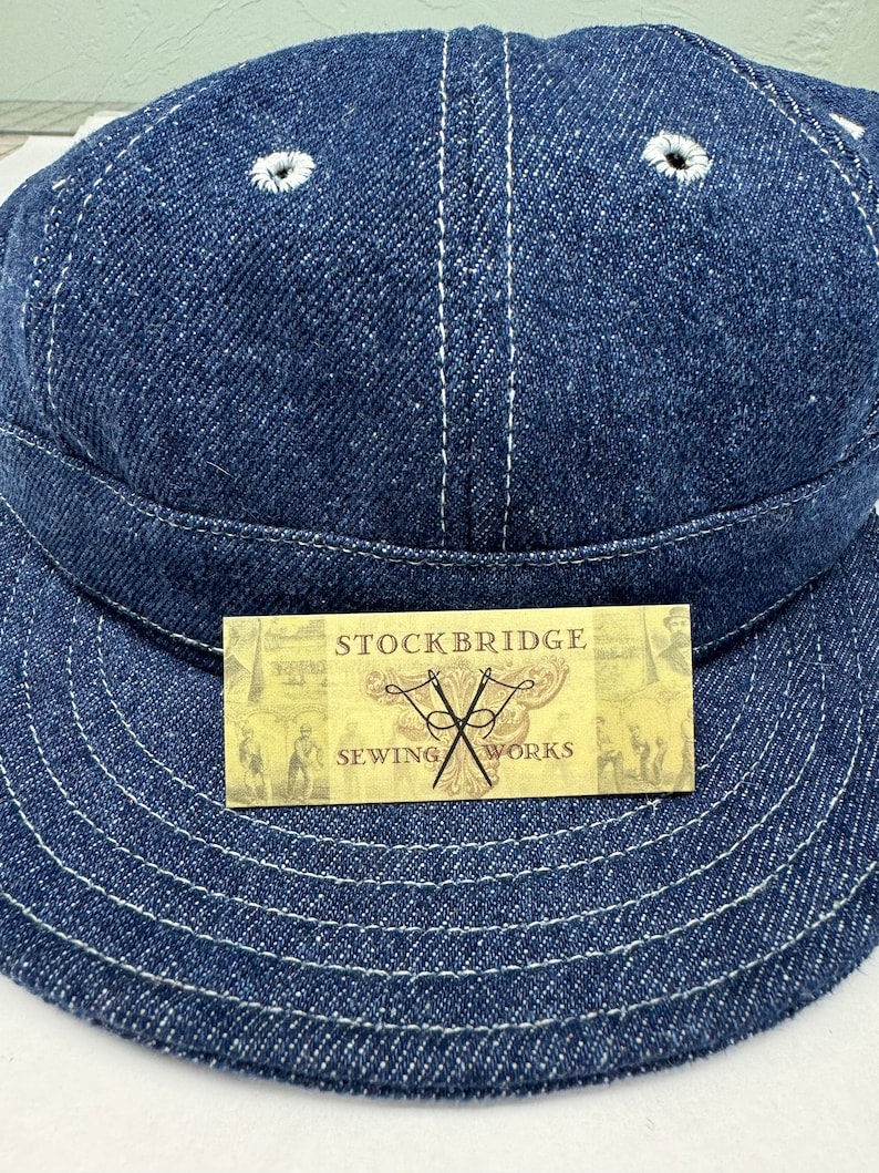 K&ouml;nnte beinhalten: Dunkelblaue Jeans-Cap mit wei&szlig;en N&auml;hten und zwei Bel&uuml;ftungs&ouml;sen. Die Cap hat einen gebogenen Schirm und ein Etikett mit der Aufschrift "STOCKBRIDGE SEWING WORKS" und einem Nadel- und Faden-Logo.