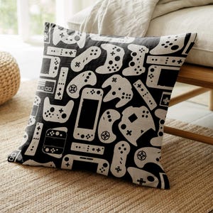 May include: Square black pillow with a white pattern of various video game controllers and handheld consoles. The pillow is made of a textured fabric and is placed on a woven rug.
