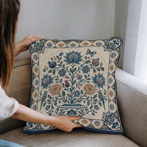 May include: A square throw pillow featuring a woven tapestry design on a cream background. The central motif displays a blue and orange floral arrangement in a vase, flanked by two green birds, a butterfly, and pomegranate-like fruits.