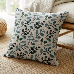 May include: Square throw pillow with a cream-colored background and a repeating pattern of blue and dark green leaves. The pillow is placed on a woven jute rug, with a wooden bench and a woven ottoman in the background.