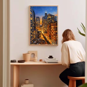 May include: A framed cityscape painting depicts a vibrant urban scene with illuminated skyscrapers under a twilight sky. The artwork is displayed on a white wall above a wooden desk with decorative items. A person is seated at the desk.