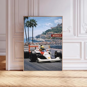 May include: Framed print of a Formula 1 race car speeding on a track in Monaco. The car is white with red and orange accents, and the driver wears a yellow helmet. The background features palm trees, the sea, and buildings.