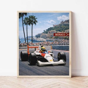 May include: Framed print of a Formula 1 race car speeding on a track in Monaco. The car is white with red and orange accents and the Marlboro logo. Palm trees, a harbor, and a sign that says "Monaco" are in the background.