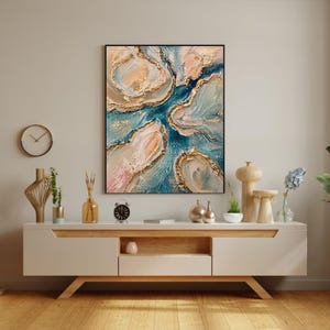 May include: Abstract art print in a black frame. The artwork features a flowing design with shades of blue, peach, and gold. The painting is displayed above a white and wood-toned cabinet with decorative items, including a clock and vases.
