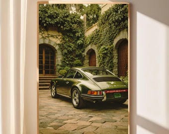 Op de afbeelding: Een ingelijste print met een vintage groene Porsche sportwagen geparkeerd op een geplaveide oprit. De auto staat voor een gebouw met met klimop begroeide muren, gewelfde deuropeningen en houten deuren. Het Porsche-logo is zichtbaar.