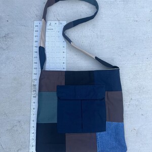 May include: A patchwork shoulder bag with a rectangular shape and a flap pocket. The bag is made of various fabric squares in shades of brown, black, blue, and denim. The strap is a combination of beige, brown, and black.