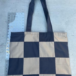 May include: A navy blue and tan checkered tote bag with matching navy blue handles. The bag is made of a sturdy fabric and is designed with a checkerboard pattern. The tote bag is shown against a concrete background.