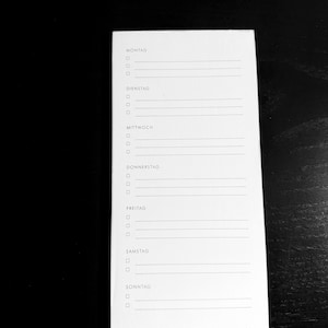 May include: A white notepad featuring a weekly planner in German. Each day, from Montag to Sonntag, has checkboxes and lines for notes. The notepad is rectangular and has a simple, minimalist design.