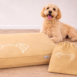 May include: A tan dog bed, drawstring bag, and a golden doodle dog. The bed and bag have a white outline of a dog's face. The dog is lying down with its tongue out. The bed is rectangular.