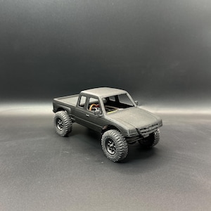 May include: A matte black scale model pickup truck featuring a detailed exterior body, a visible steering wheel inside the cabin, and four rugged, textured off-road tires. The vehicle has a standard truck bed and a front grille design.