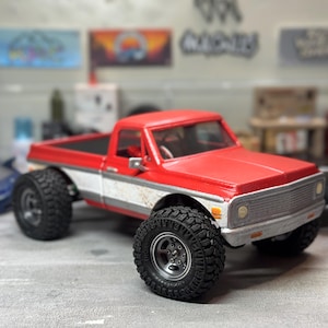 May include: A red and white toy pickup truck with large black tires. The truck has a silver grill and a black bed. The toy truck is sitting on a gray surface.