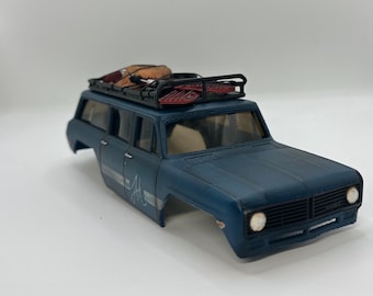 May include: A matte blue die-cast station wagon model featuring a black roof rack loaded with red snowshoes and a brown gear bag. The vehicle body displays a white script logo on the side, a black front grille, and circular headlights.