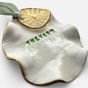 May include: A white ceramic ornament with a gold rim and the name "TREVOR" in green lettering. A dried citrus slice is attached to the top with a green ribbon. The ornament has a wavy, organic shape.