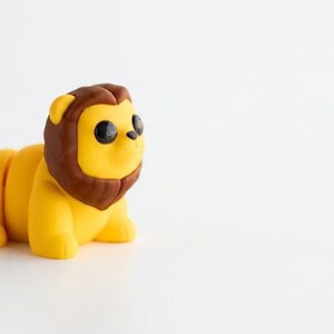 May include: A small, stylised lion figurine with a bright yellow body and a dark brown mane. The lion has large, round, black eyes, a small black nose, and a brown tuft at the end of its tail. The figure is posed in a standing position.