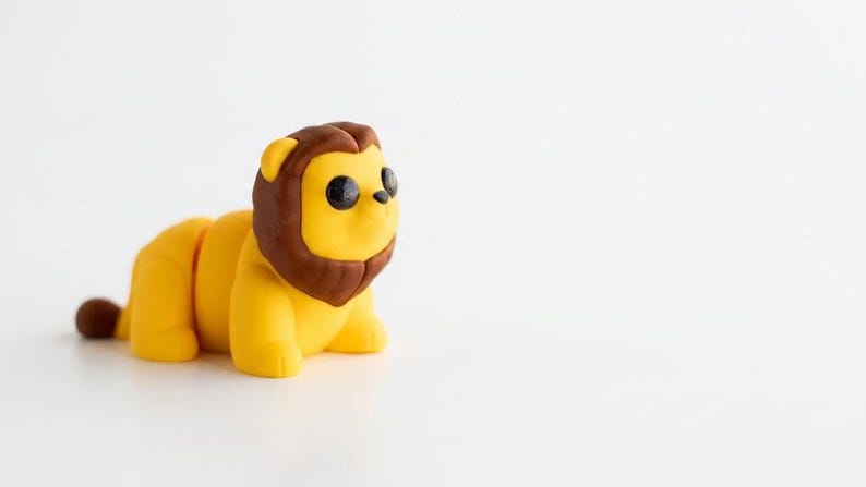 May include: A small, stylised lion figurine with a bright yellow body and a dark brown mane. The lion has large, round, black eyes, a small black nose, and a brown tuft at the end of its tail. The figure is posed in a standing position.