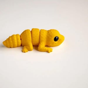 May include: A bright yellow articulated lizard figurine with a textured, bumpy surface and smooth black eyes. The creature features a segmented body design that allows for movement, with four small legs and a tapered tail, presented in a solid colour.