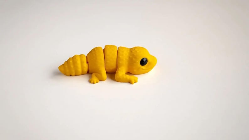 May include: A bright yellow articulated lizard figurine with a textured, bumpy surface and smooth black eyes. The creature features a segmented body design that allows for movement, with four small legs and a tapered tail, presented in a solid colour.