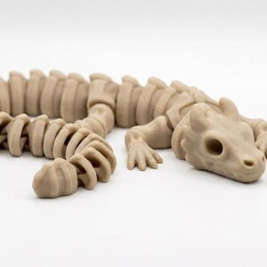 May include: A light tan, articulated dragon figurine featuring a segmented, flexible body structure with a skeletal appearance. The creature has a detailed head with hollow eye sockets, small horns, and four limbs with clawed feet, all in a matte finish.