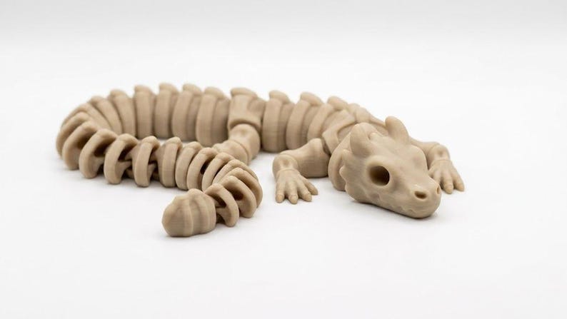 May include: A light tan, articulated dragon figurine featuring a segmented, flexible body structure with a skeletal appearance. The creature has a detailed head with hollow eye sockets, small horns, and four limbs with clawed feet, all in a matte finish.