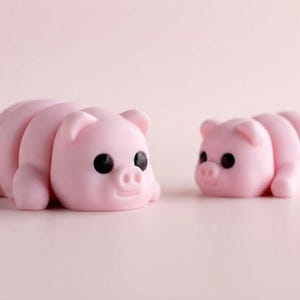 May include: Two small, light pink pig figurines with smooth, rounded bodies and segmented backs. Each pig features large, solid black circular eyes, small upright ears, and a snout with two nostrils, positioned facing each other on a flat surface.