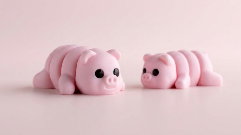 May include: Two small, light pink pig figurines with smooth, rounded bodies and segmented backs. Each pig features large, solid black circular eyes, small upright ears, and a snout with two nostrils, positioned facing each other on a flat surface.