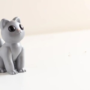 May include: A small, smooth, light grey cat figurine with large, round, glossy black eyes and a tiny pink nose. The cat is depicted in a seated position, looking upward with pointed ears and a short, upright tail, featuring a solid matte finish.
