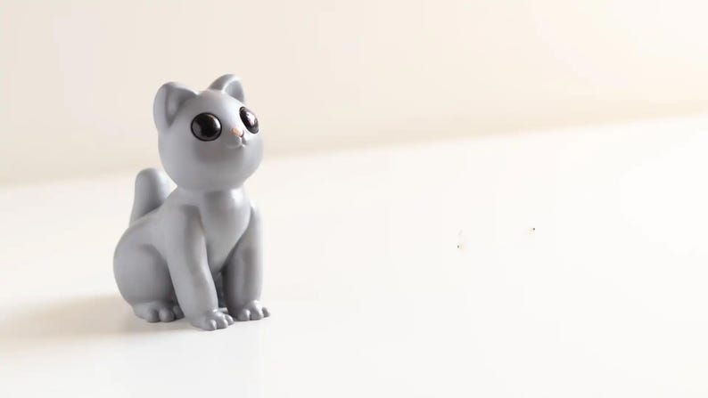 May include: A small, smooth, light grey cat figurine with large, round, glossy black eyes and a tiny pink nose. The cat is depicted in a seated position, looking upward with pointed ears and a short, upright tail, featuring a solid matte finish.