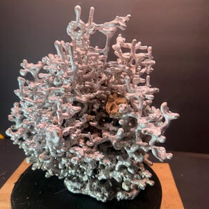 May include: A detailed, silver-colored sculpture with a complex, organic structure, resembling a miniature landscape or a cast of a natural form. The sculpture is mounted on a small, round, black base, and sits on a wooden surface.
