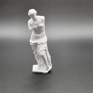 May include: A white, classical statue of a woman, missing both arms. The figure is draped in fabric and stands on a rectangular base. The statue is a representation of classical art.