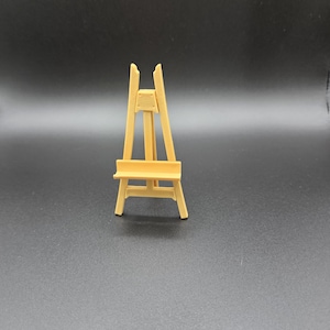 May include: A small, light brown wooden easel, designed to hold a small canvas or artwork. The easel has a three-legged design with a support shelf and a vertical back for stability. The wood has a natural, unpainted finish.