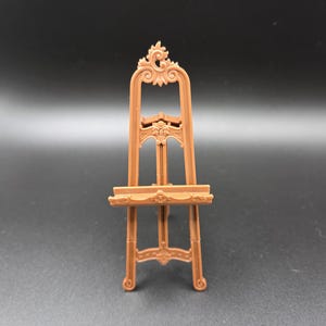 May include: A small, brown decorative easel with ornate scrollwork at the top and on the shelf. The miniature art stand has a three-legged base and is designed to hold a small canvas or artwork.