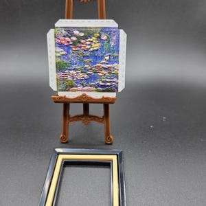 May include: Miniature brown easel displaying a painting of water lilies in a blue pond, with pink, green, and yellow accents. A black and tan picture frame sits below the easel. The painting is a miniature replica of a famous artwork.