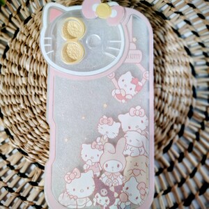 May include: Pink and clear phone case featuring a Hello Kitty design. The case has a pink border and a clear center with glitter and Hello Kitty characters. The top of the case has a Hello Kitty head design with a pink bow.