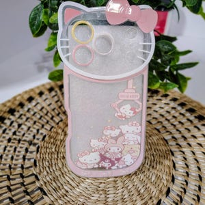 May include: A pink and clear phone case featuring a Hello Kitty design. The case has a bow at the top and a pink border. The case has a clear section with Hello Kitty characters and the words "Hello Kitty".