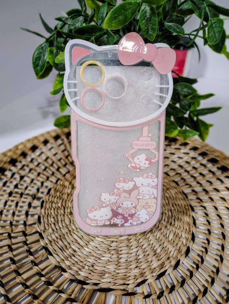 May include: A pink and clear phone case featuring a Hello Kitty design. The case has a bow at the top and a pink border. The case has a clear section with Hello Kitty characters and the words "Hello Kitty".