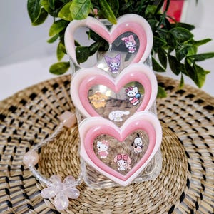 May include: A clear phone case with a pink heart-shaped design. The case features multiple cartoon characters inside the hearts. A beaded bracelet with a flower charm is next to the phone case. The background is a woven mat and green plant.