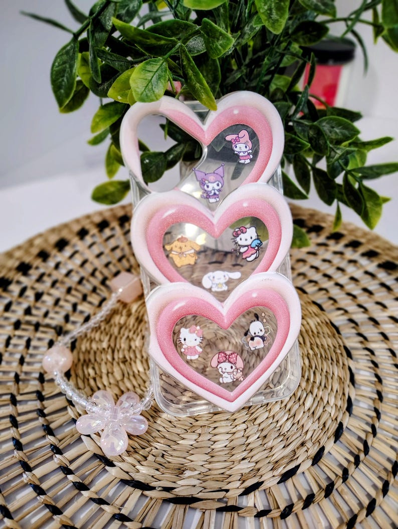 May include: A clear phone case with a pink heart-shaped design. The case features multiple cartoon characters inside the hearts. A beaded bracelet with a flower charm is next to the phone case. The background is a woven mat and green plant.