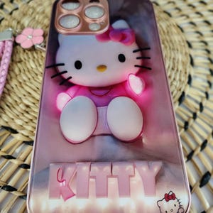 May include: Pink phone case with a 3D Hello Kitty design. The case features a pink bow, black whiskers, and the word "KITTY" in raised letters. A small Hello Kitty graphic is in the bottom right corner. The case is for a smartphone.
