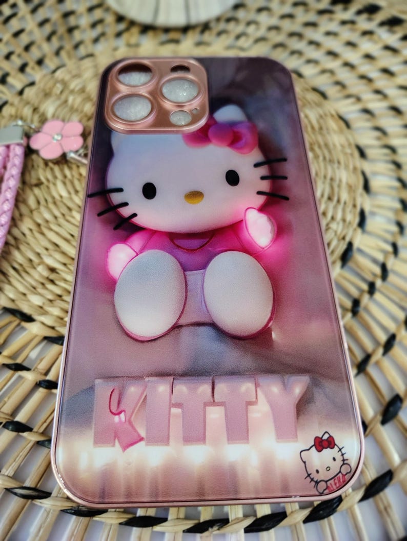 May include: Pink phone case with a 3D Hello Kitty design. The case features a pink bow, black whiskers, and the word "KITTY" in raised letters. A small Hello Kitty graphic is in the bottom right corner. The case is for a smartphone.