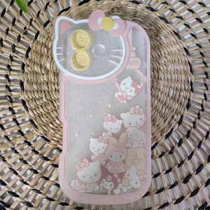 May include: Pink phone case featuring a Hello Kitty design. The case has a clear center with floating glitter and Hello Kitty characters. The top has a Hello Kitty head shape with two gold-colored circles. The case is on a woven background.