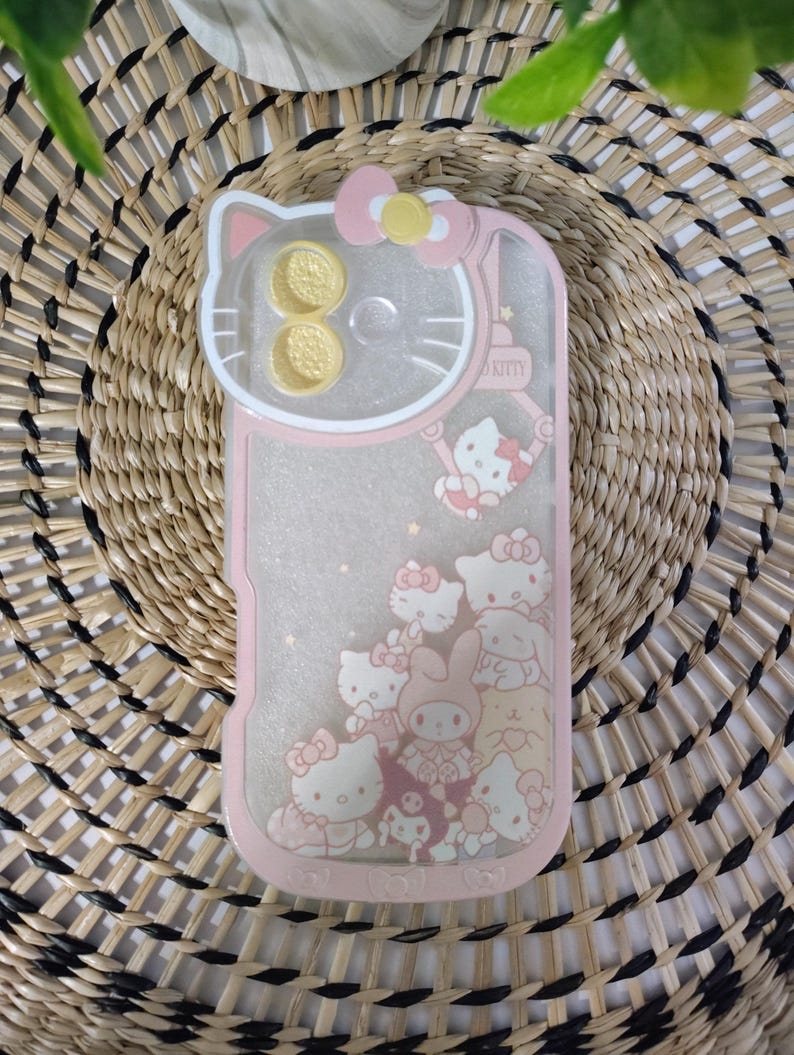 May include: Pink phone case featuring a Hello Kitty design. The case has a clear center with floating glitter and Hello Kitty characters. The top has a Hello Kitty head shape with two gold-colored circles. The case is on a woven background.