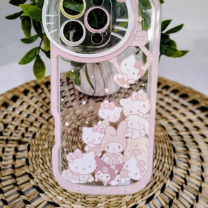 May include: Pink and clear phone case featuring a Hello Kitty design. The case has a pink border and a clear center with floating Hello Kitty characters. The top of the case has a pink bow and the word "Kitty".