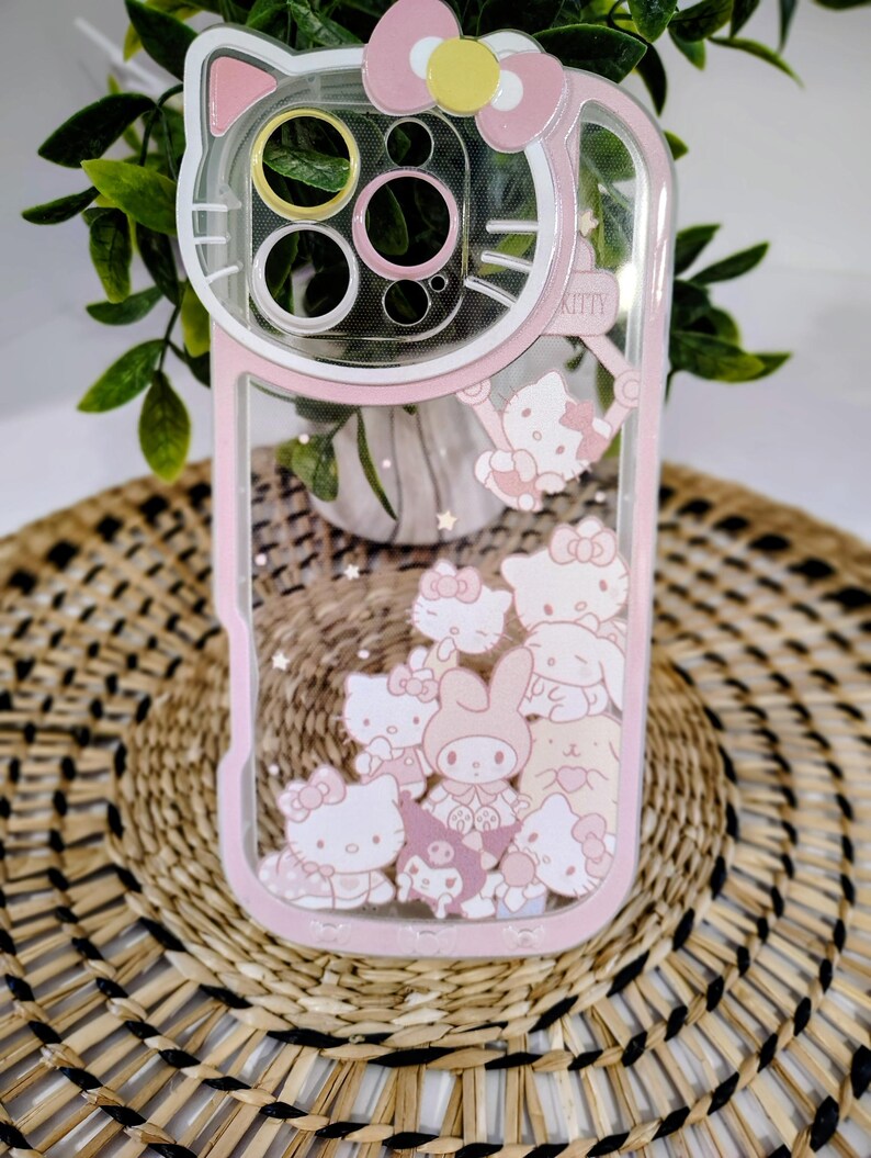 May include: Pink and clear phone case featuring a Hello Kitty design. The case has a pink border and a clear center with floating Hello Kitty characters. The top of the case has a pink bow and the word "Kitty".