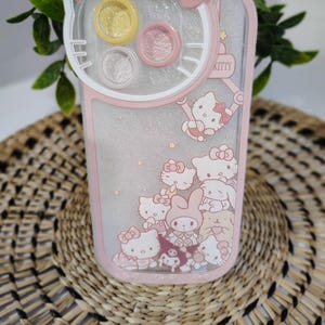 K&ouml;nnte beinhalten: Rosa-wei&szlig;es Handyh&uuml;lle mit Hello Kitty-Design. Die H&uuml;lle hat ein klares, glitzerndes Innere mit mehreren Hello Kitty-Figuren. Die Oberseite der H&uuml;lle hat eine Schleife und drei kreisf&ouml;rmige F&auml;cher. Das Wort "KITTY" ist sichtbar.