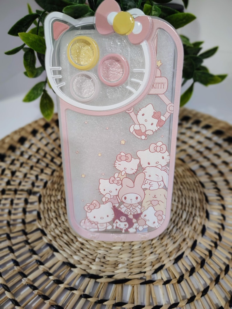 K&ouml;nnte beinhalten: Rosa-wei&szlig;es Handyh&uuml;lle mit Hello Kitty-Design. Die H&uuml;lle hat ein klares, glitzerndes Innere mit mehreren Hello Kitty-Figuren. Die Oberseite der H&uuml;lle hat eine Schleife und drei kreisf&ouml;rmige F&auml;cher. Das Wort "KITTY" ist sichtbar.