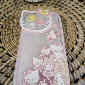 K&ouml;nnte beinhalten: Eine rosa und transparente Handyh&uuml;lle mit Hello Kitty und ihren Freunden. Die H&uuml;lle hat einen rosa Rand und eine transparente Mitte mit Glitzer und Charakterillustrationen. Die Oberseite der H&uuml;lle hat die Form eines Hello Kitty-Kopfes.