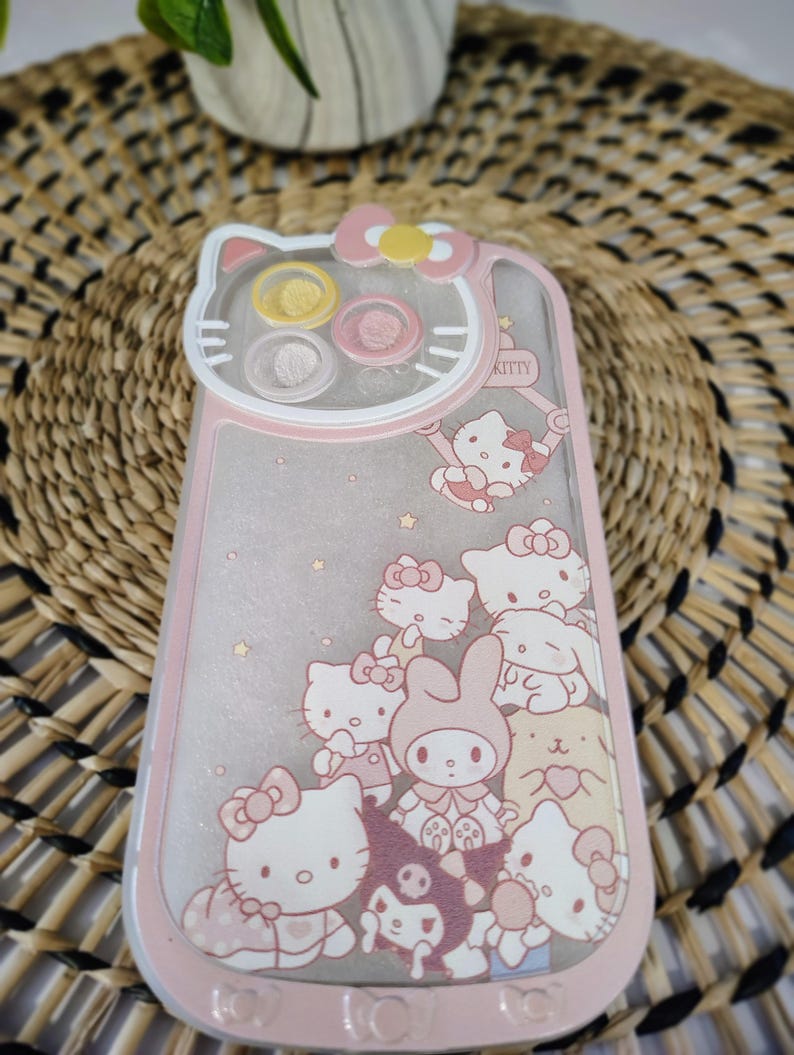 K&ouml;nnte beinhalten: Eine rosa und transparente Handyh&uuml;lle mit Hello Kitty und ihren Freunden. Die H&uuml;lle hat einen rosa Rand und eine transparente Mitte mit Glitzer und Charakterillustrationen. Die Oberseite der H&uuml;lle hat die Form eines Hello Kitty-Kopfes.